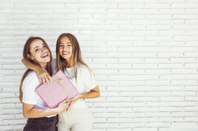 dos-amigas-felices-regalo-cumpleanos-frente-pared-ladrillo_23-2147888251